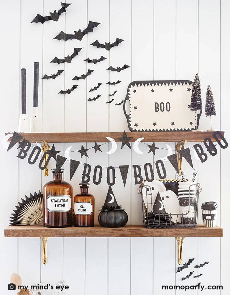 Floating shelves decorated with modern Halloween decorations including My Mind's Eye vintage Halloween boo banner, witching hour witch party hats, paper cutout bats, Boo reusable bamboo tray and a boo basket filled with ghost shaped plates, napkins, and cups, with vintage bottle that look like potion.