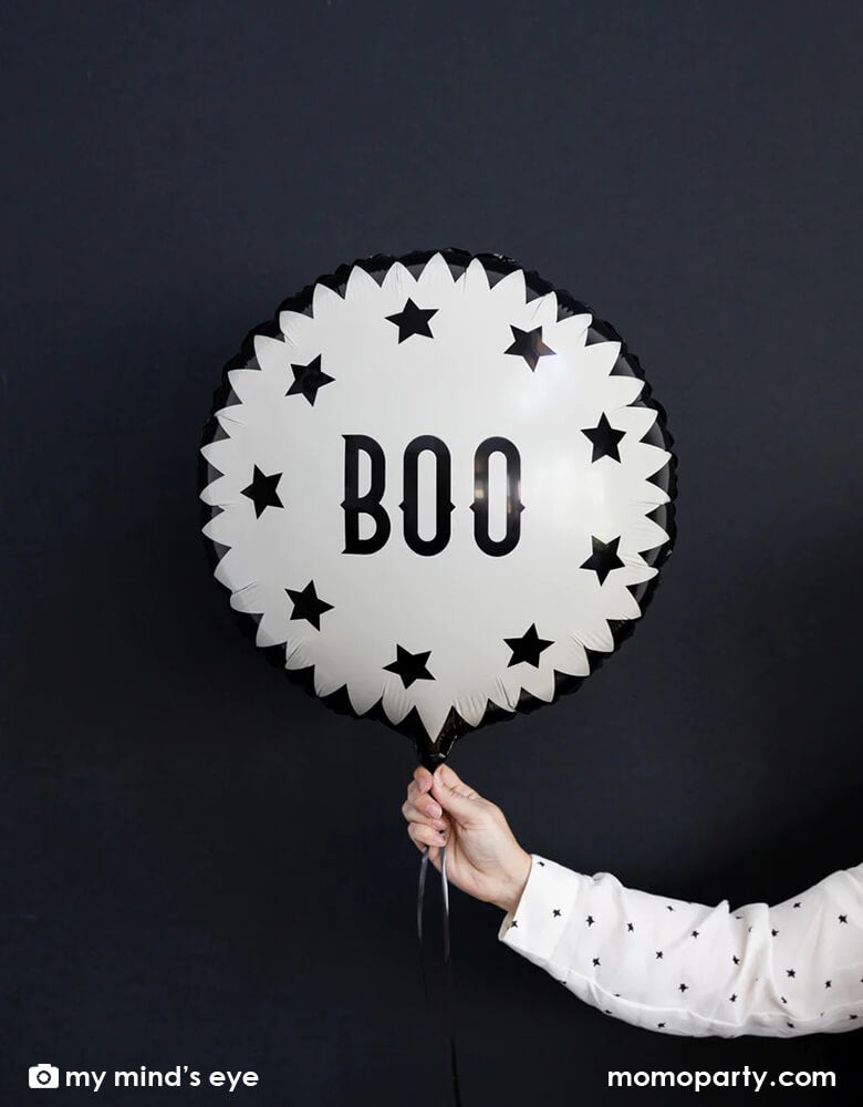 A woman's hand with white polka dot shirt holding My Mind's Eye Vintage Halloween Boo Mylar Balloon against black background creating a spooky vibe for a Halloween celebration 