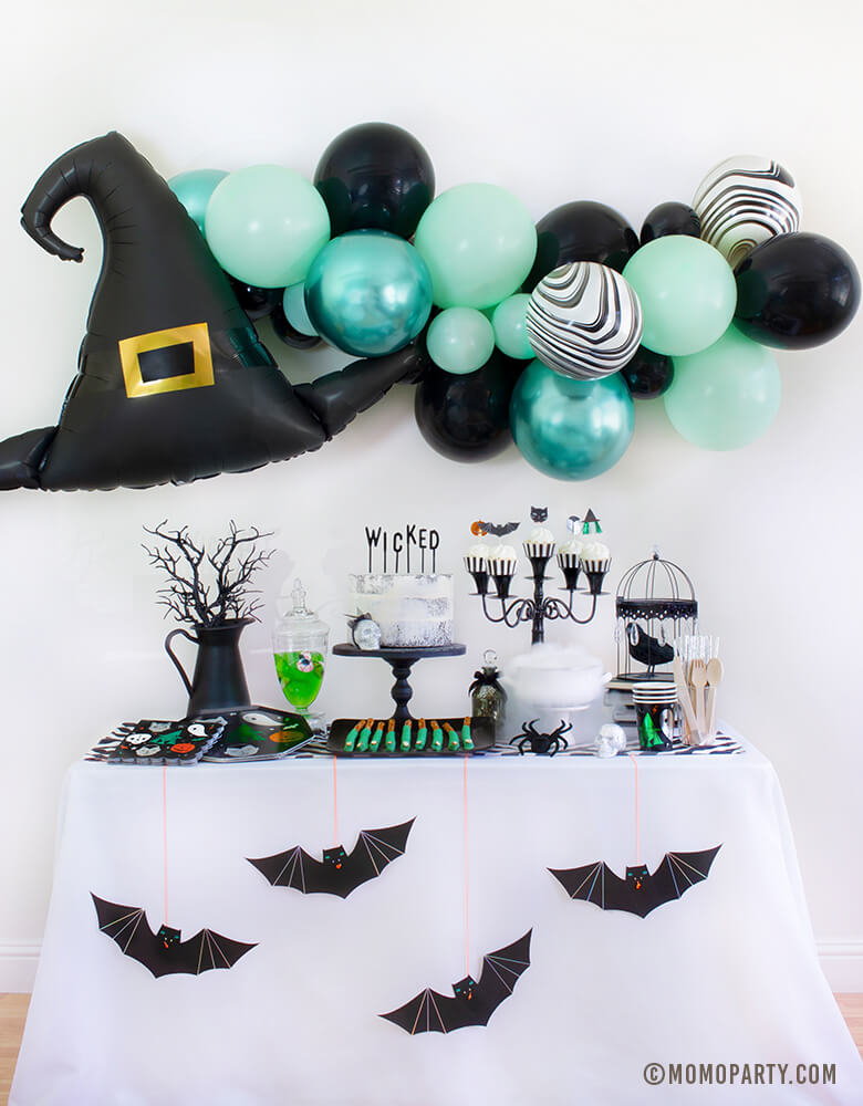 Momo Party 2020 Witch Please themed Halloween Party at home inspiration look with Anagram Witch Hat Satin Foil Balloon and pastel Green toned Latex Balloon Garland as wall decoration backdrop. Table set up with Meri Meri Halloween Motif Dinner Plates, napkins and cups, cake with letter board cake topper spelled of "Wicked", smoking witch cauldron made of dry ice, Apple jelly in with eyeball candy in a jar, witch fingers pretzels in a black plate for dessert, Meri Meri Hanging Bats in front of the table 