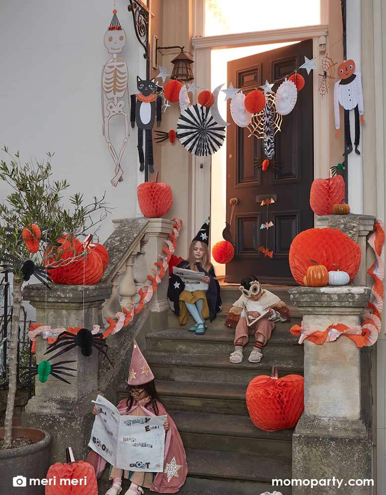 A peak of a doorstep decorated Classic Halloween decorations in orange and black with kids dressed-up for Halloween. The Halloween decorations featured are Meri Meri's Halloween Honeycomb party fan banner and hanging paper decorations of a skeleton, a pumpkin person and a black cat. The stairs were topped with Meri Meri's giant pumpkin honeycomb paper decorations, a perfect inspiration for kid's not-so-scary Halloween home decorations