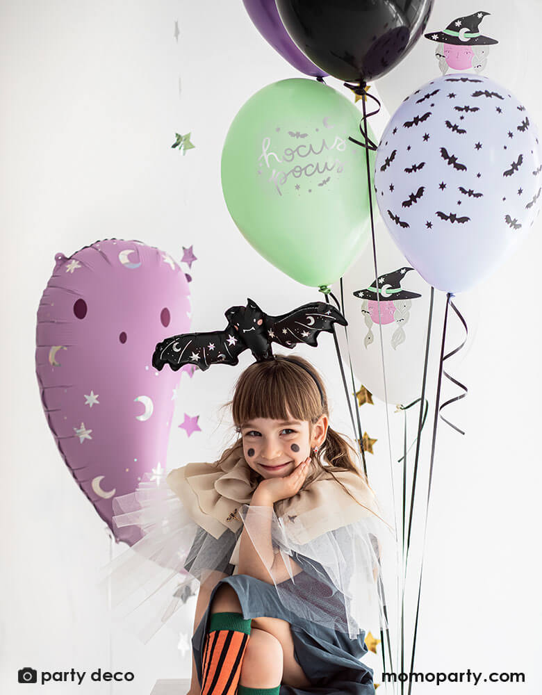 A little girl with Halloween themed balloons including a pink ghost shaped foil balloons and latex balloons in mint, purple, and black with witch illustrations
