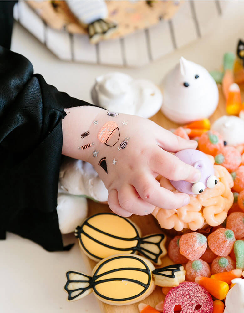 A girl in a witch costume with daydream society's hocus pocus temporary tattoos on her hand reaches out to a bowl of halloween themed treats and candies in a kids Halloween party