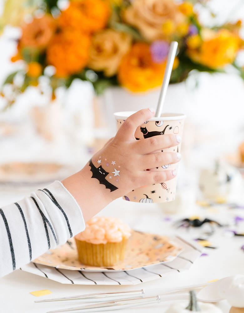 A boy in a mummy costume with daydream society's hocus pocus temporary tattoos on his hand holding a party cup in a fun kid's Halloween party full of pretty fall flower arrangment and delicious treats