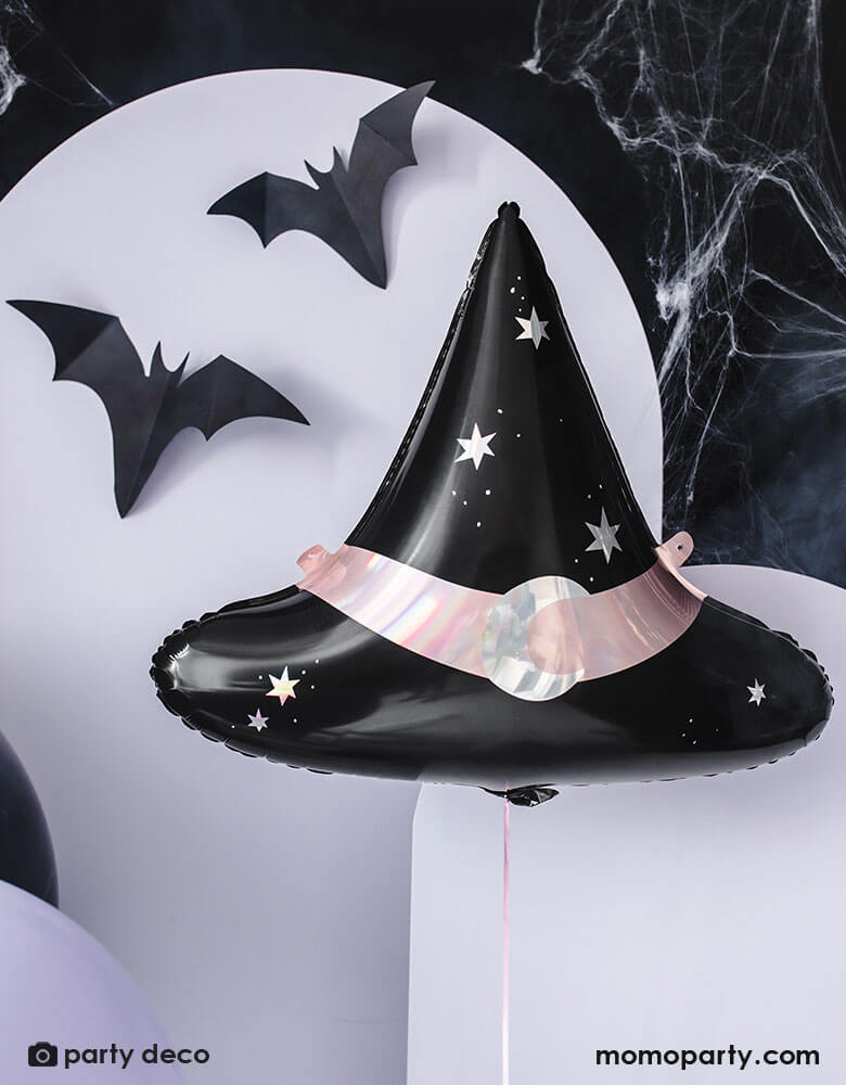 Momo Party's 23.5 x 19 inches Hocus Pocus witch hat shaped foil balloon by Party Deco hung next to some wooden panel party backdrop in lilac color which is adorned with bat shaped paper decoration and spiderweb, making this a perfect look for a fun Halloween party this spooky season.