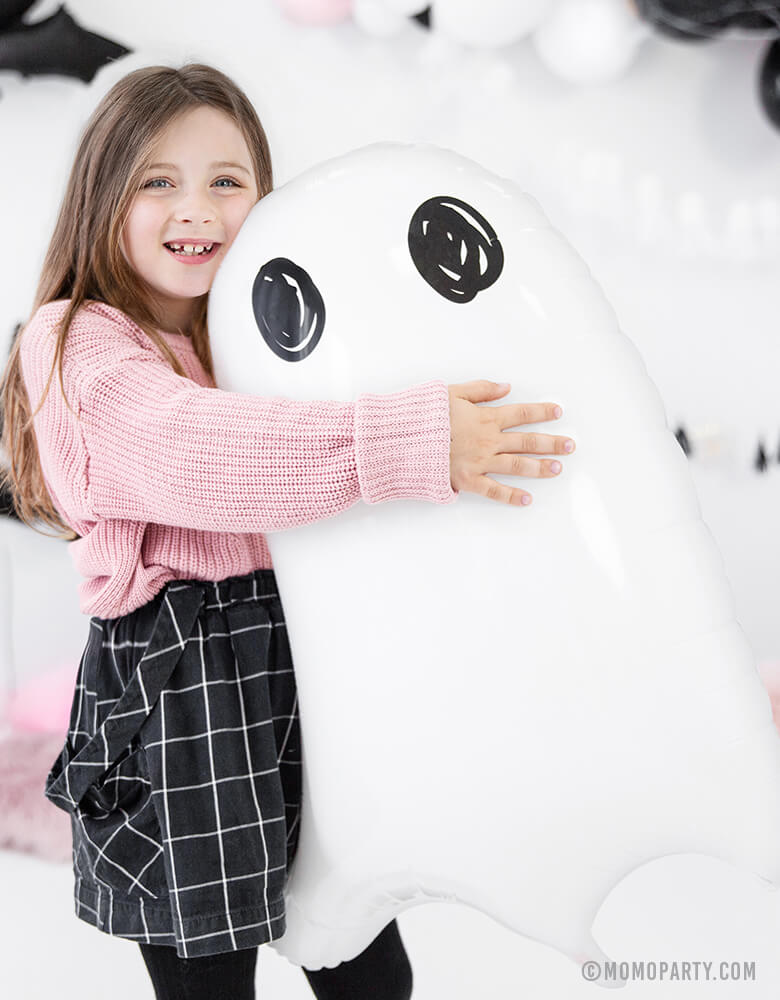 A girl wearing a pink sweater happily holding a Party Deco Halloween Ghost Foil Mylar Balloon. This cute clean modern look Ghost foil balloon is perfect for a kid-friendly modern spooky halloween party, trick-or-treating halloween party, nightmare before Christmas party, witch themed party and all halloween related celebrations