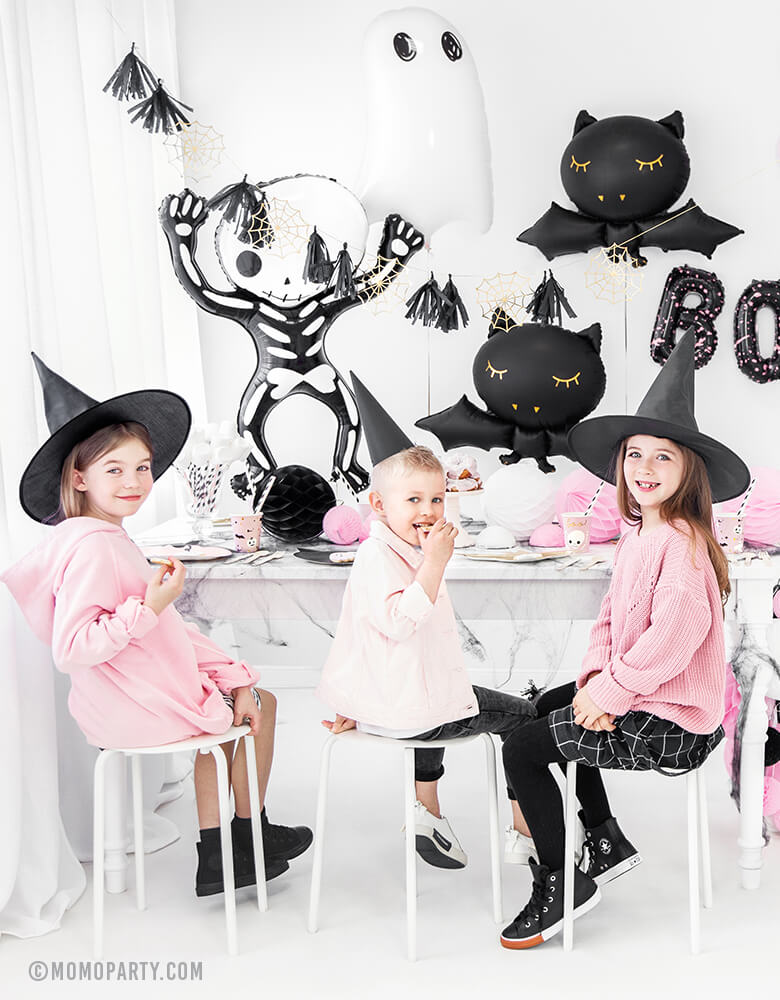  boy and girls wearing witch hats, seating in front of party table, celebrating a Pink Halloween. Room decorated with Party Deco Halloween Skeleton shaped Foil Mylar Balloon, Halloween Ghost Foil Mylar Balloon, Black bat foil balloon, and golden spiderwebs and black tassels garland. These halloween balloons also perfect for Kid-Friendly modern spooky halloween party, trick-or-treating halloween party, nightmare before christmas party, witch themed party and all halloween related celebrations