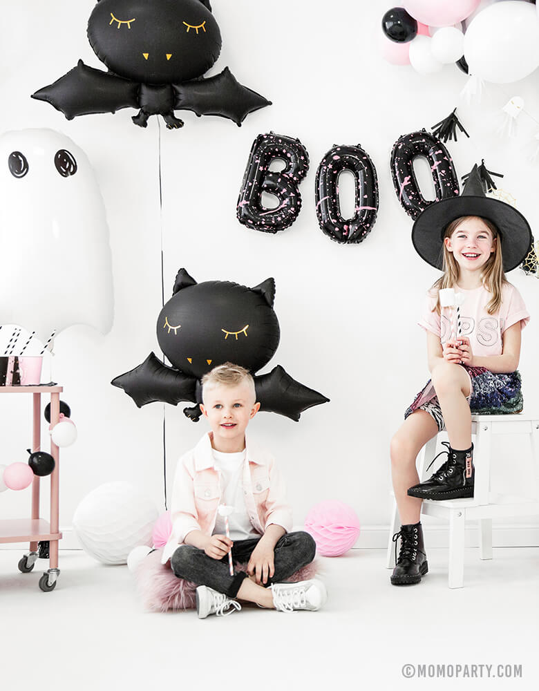 a boy and a girl who wearing witch hat, seating in a room decorated with Party Deco Halloween Skeleton shaped Foil Mylar Balloon, Halloween Ghost Foil Mylar Balloon, Black bat foil balloon, and golden spiderwebs and black tassels garland, celebrating a Pink Halloween. These halloween balloons also perfect for Kid-Friendly modern spooky halloween party, trick-or-treating halloween party, nightmare before christmas party, witch themed party and all halloween related celebrations