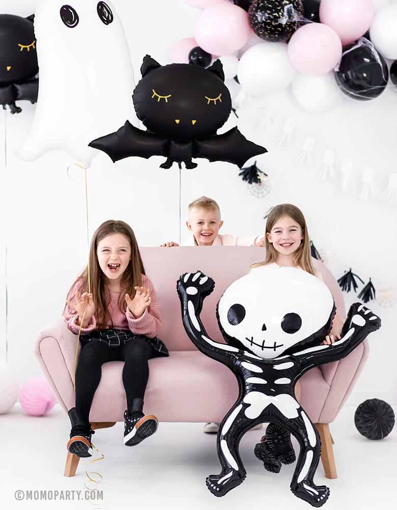 boy and girls seating on the sofa and holding a Party Deco Halloween Skeleton shaped Foil Mylar Balloon, with a Halloween Ghost Foil Mylar Balloon, and Black bat foil balloon, pink white and black mixed balloon garland on the wall, celebrating a Kid-Friendly modern spooky halloween party, trick-or-treating halloween party, nightmare before christmas party, witch themed party and all halloween related celebrations