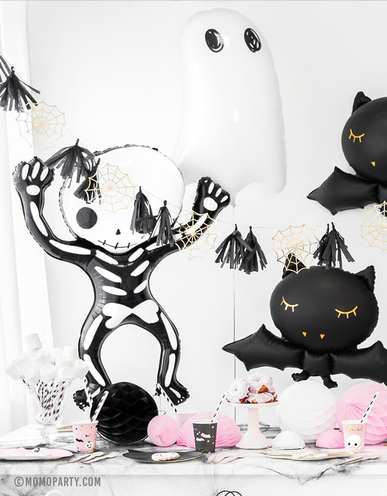 Pastel Pink Halloween party table full of halloween tablewares, decorated with Party Deco Halloween Skeleton shaped Foil Mylar Balloon, Halloween Ghost Foil Mylar Balloon, Black bat foil balloon, and golden spiderwebs and black tassels garland. These halloween balloons also perfect for Kid-Friendly modern spooky halloween party, hocus pocus party trick-or-treating party, nightmare before christmas party, witch themed party and all halloween related celebrations