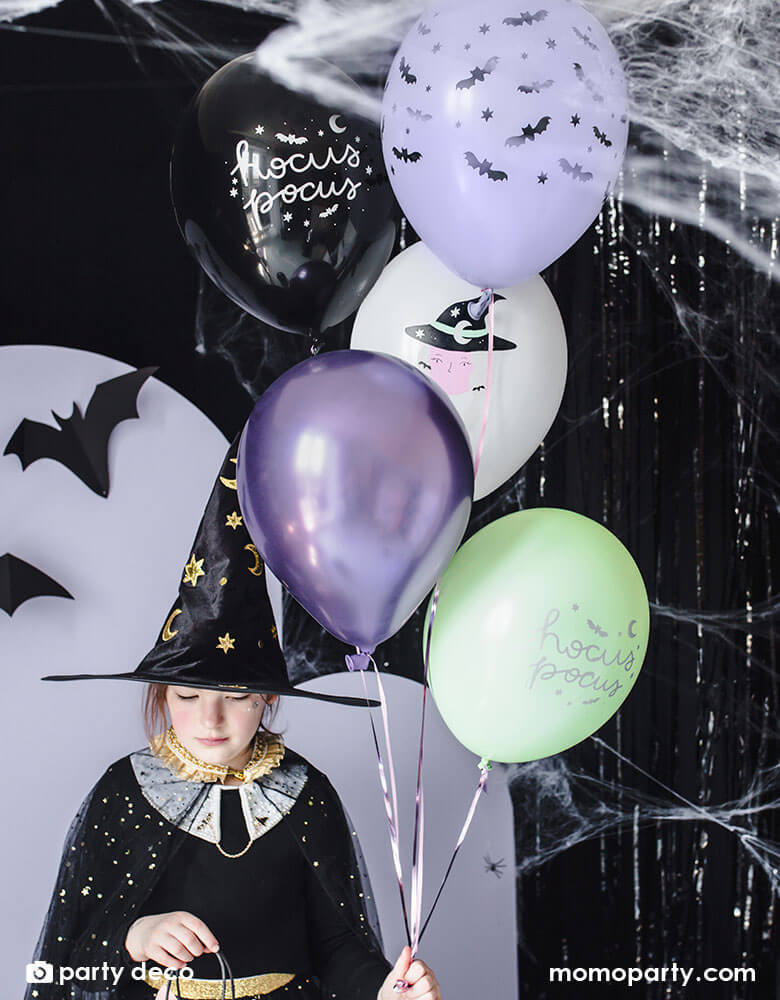 A witch Hocus Pocus themed Halloween party set up with black and purple themed color backdrop with a little girl dressed up in Witch cap and hat standing next to a latex balloon mix of Party Deco 11" Witch Balloon Mix comes with 6 assorted latex balloons with witch & bats illustrations and hocus pocus words in a combination of black, purple, mint, and lilac colors