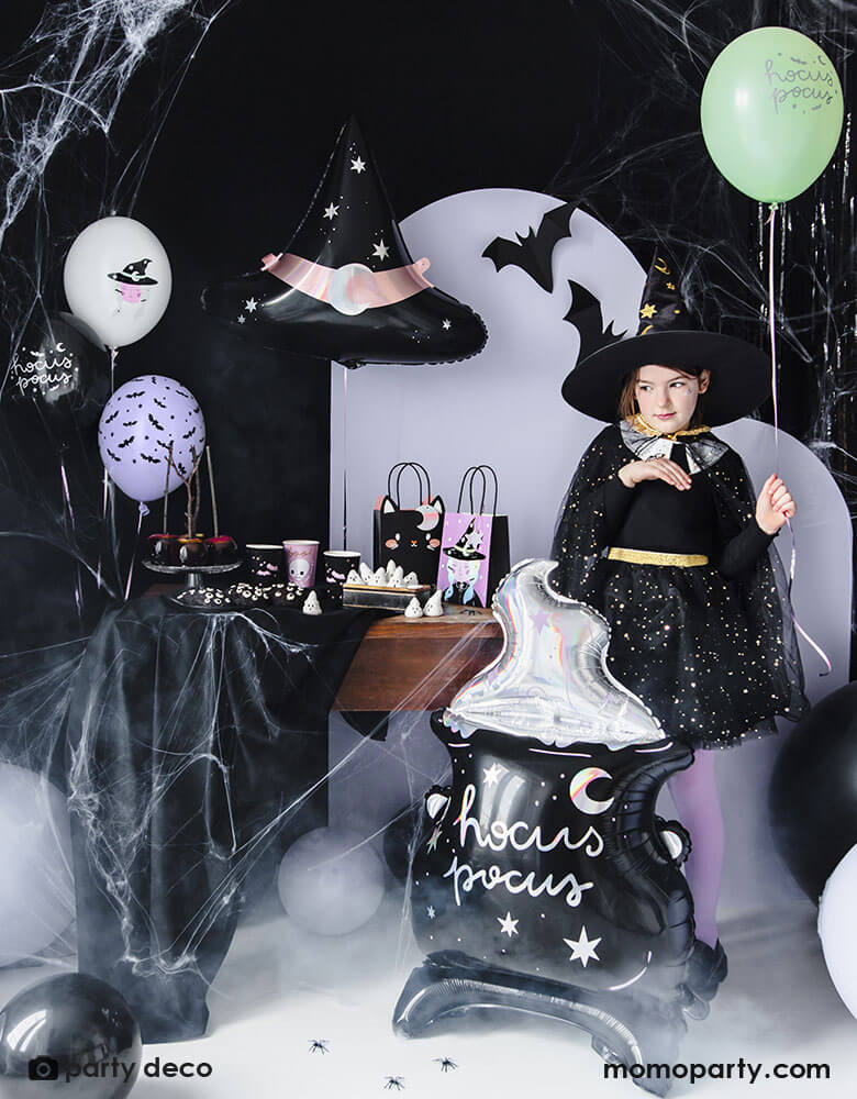 A witch Hocus Pocus themed Halloween party set up with black and purple themed color backdrop with witch hat foil balloon and Hocus Pocus Cauldron standing balloon with a little girl dressed up in Witch cap and hat standing next to a latex balloon mix of Party Deco 11" Witch Balloon Mix comes with 6 assorted latex balloons with witch & bats illustrations and hocus pocus words in a combination of black, purple, mint, and lilac colors