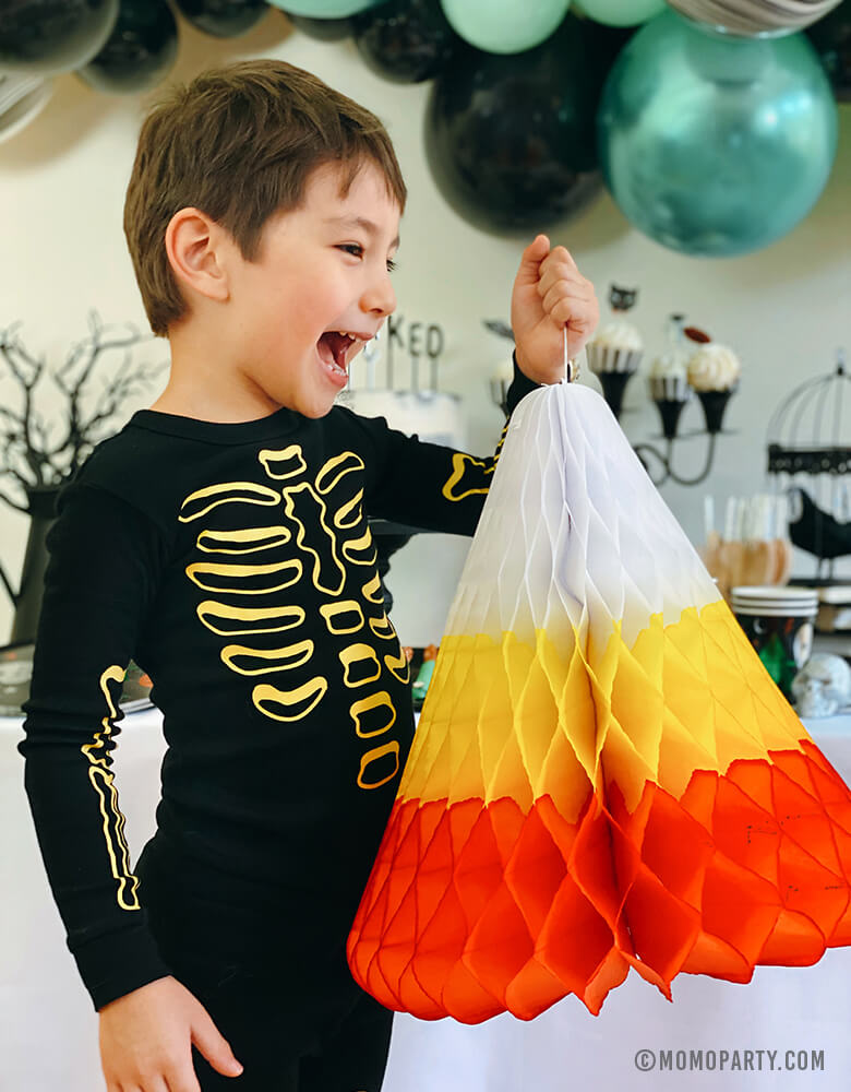 boy wearing skeleton pjs, happily holding a huge paper made candy corn honeycomb decoration in front of a witch themed halloween birthday party with chrome green, black, pastel matt mint color mixed balloon garland. This huge Hand-crafted and hand-dyed Candy Corn in the traditional white, yellow, and orange stripes, unique fun party decoration for sweeten your little one's halloween party!