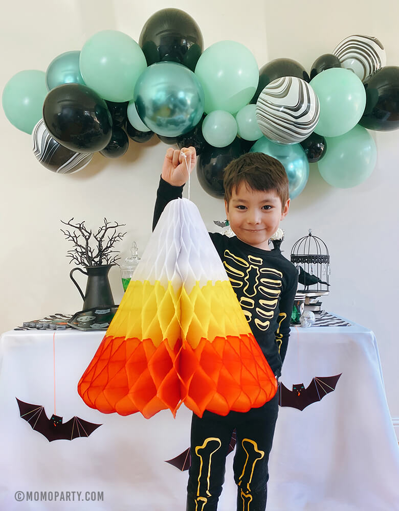 A boy holding a huge candy corn honeycomb decoration in front of halloween party table, there are meri meri hanging bats in the front of the dessert table, chrome green, black, pastel matt mint mixed latex balloon garland as backdrop. The huge Hand-crafted and hand-dyed Candy Corn made in the traditional white, yellow, and orange stripes, super cool decoration for A Modern trick-or-treating halloween party, nightmare before christmas party, witch themed party and all halloween related celebrations