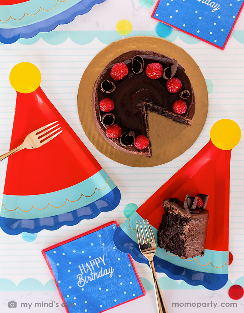 A festive birthday party table with a chocolate birthday cake, filled with cheerful confetti in blue, red and yellow and My Mind's Eye blue Happy Birthday napkins with blue birthday hat shaped party plates - a perfect size for serving birthday cake - great modern and simple ideas for a boy's birthday celebration.