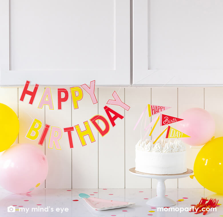 A simple and modern at-home birthday party decoration set up for a girl's birthday party at the kitchen counter space with pink and yellow birthday party balloons, My Mind's Eye's pink birthday Banner in cute colors of pink coral, yellow and red red, yellow, with a birthday cake topped with My Mind's Eye Happy birthday pink pennant cake toppers with cheerful ribbons in darling pink and peach. Perfect for a girly themed birthday party!