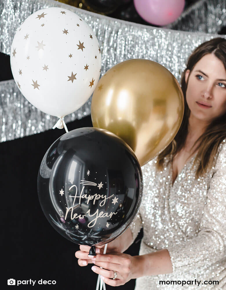 A lady in sequin party dress holding Momo Party's Happy New Year Latex Balloon Mix, set of 6 by Party Deco in the color of black, white and gold with Happy New Year message on it, a perfect decoration for a New Years Party celebration!