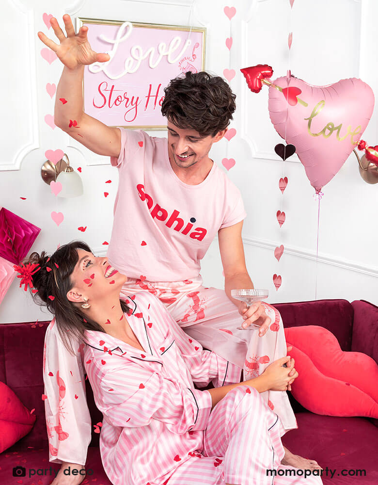 A couple in their pink pajamas celebrating Valentine's Day sprinkling heart shaped tissue confetti in pink and red, with a pink heart shaped foil balloon with an arrow crossing it. They're showing love to each other on a bold red velvet couch with lips shaped pillow, creating a fun yet romantic scene. 