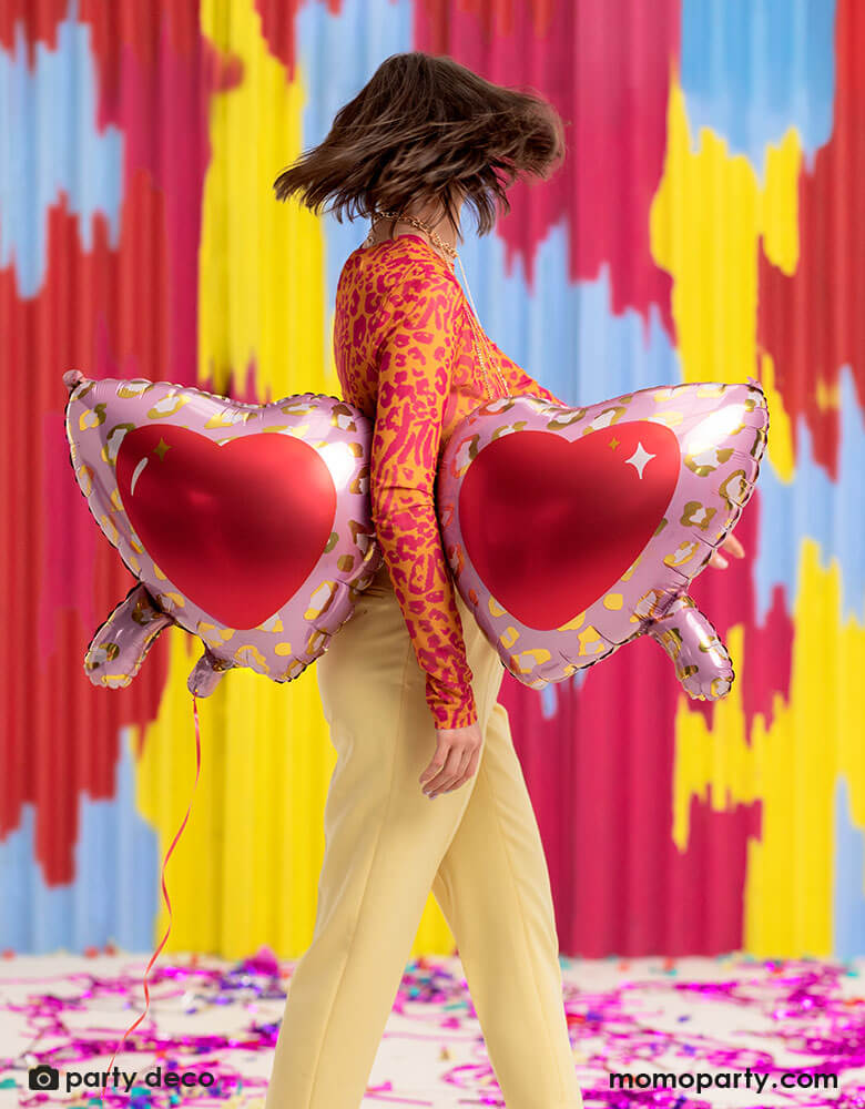 A young lady in her leopard shirt holding Momo Party's 30 inch heart shaped sunglasses foil balloon by Party Deco, With animal print design around the glasses in pink and red colors in her arms, standing in front of a colorful bright wall, celebrating love for Valentine's Day.