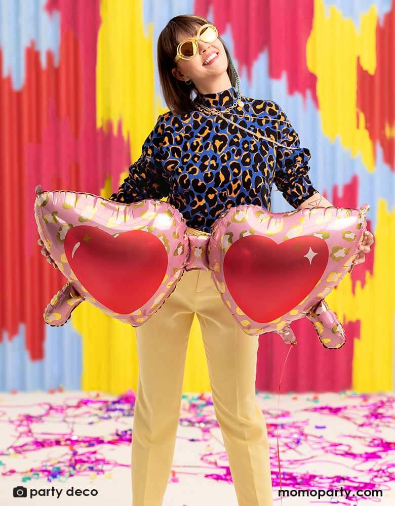A young lady in her leopard shirt holding Momo Party's 30 inch heart shaped sunglasses foil balloon by Party Deco, With animal print design around the glasses in pink and red colors in her arms, standing in front of a colorful bright wall, celebrating love for Valentine's Day.