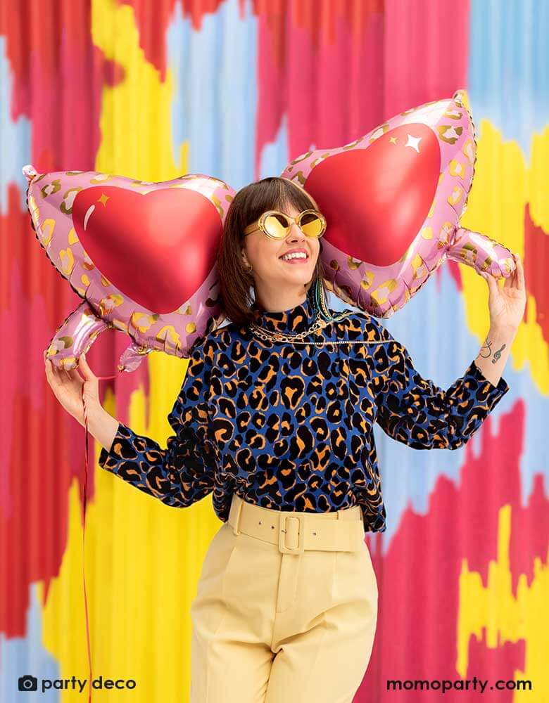 A young lady in her leopard shirt holding  Momo Party's 30 inch heart shaped sunglasses foil balloon by Party Deco, With animal print design around the glasses in pink and red colors on her shoulder, standing in front of a colorful bright wall, celebrating love  for Valentine's Day.