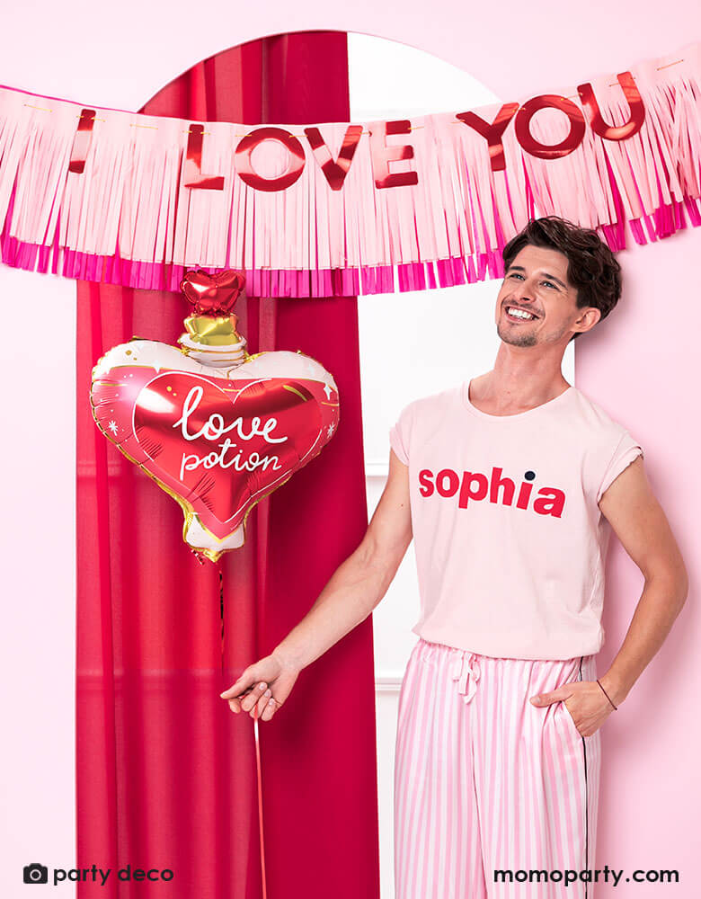 A man dressed in pink pajama holding Momo Party's love potion shaped foil balloon by Party Deco, standing in front of a door decorated with pink tissue-fringe banner with red type message of "I love you" - a perfect way to express your love on Valentine's Day.