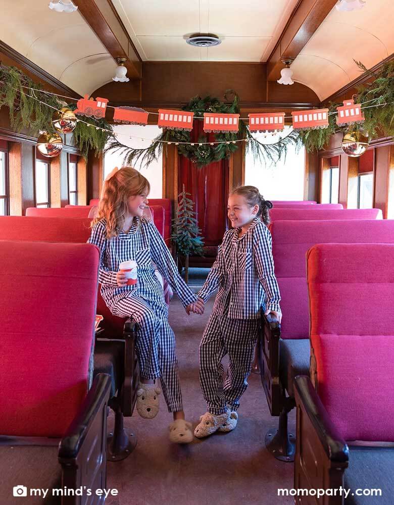 Two little girls in their Holiday pajamas holding North Pole Express red 8 oz to-go cups by My Mind's Eye sitting on a vintage Polar Express train cabin decorated with Momo Party's North Pole Express train and bell garland set, a perfect idea for this Holiday season's celebration with family and kids.