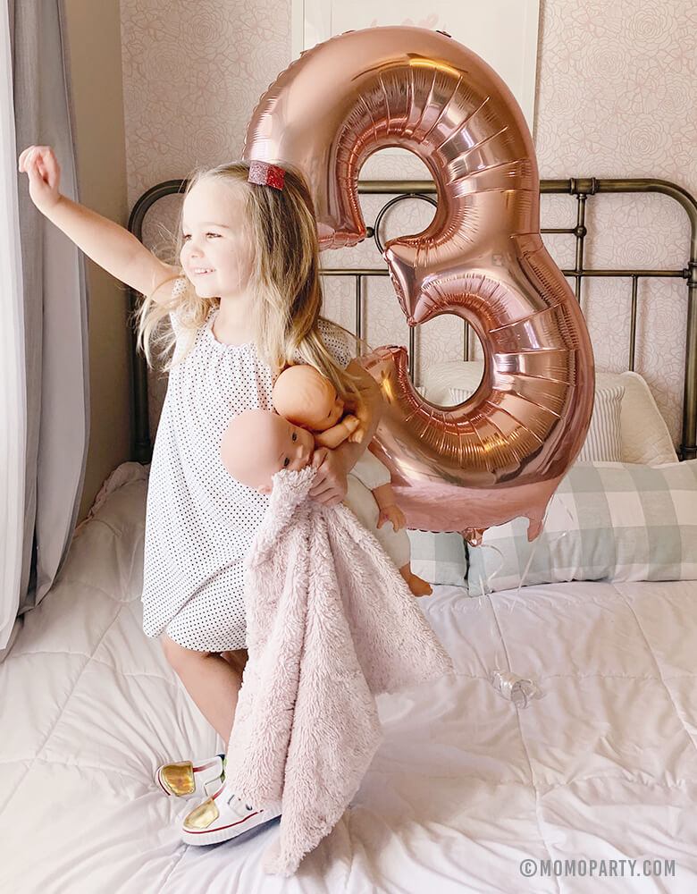 A blond girl holding a baby doll and a Large Rose Gold Number 3 Foil Mylar Balloon standing on her bed looking out window at her 3 years birthday