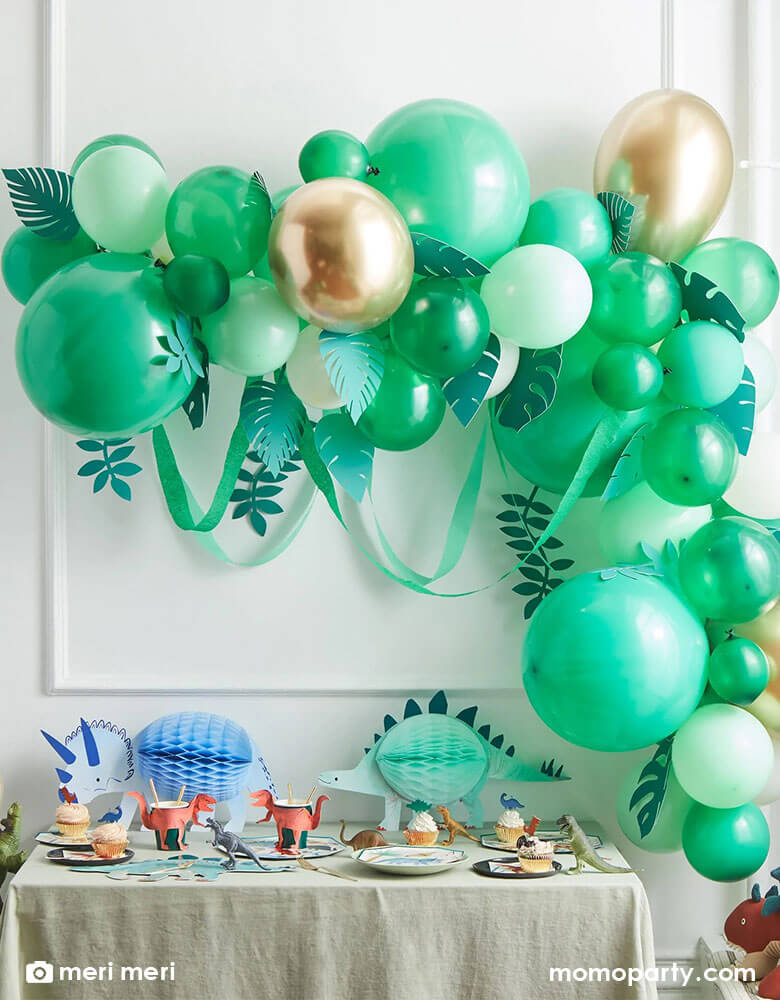 A kid's dinosaur themed birthday party with a wall decorated with Meri Meri's leafy balloon arch including 44 balloons, including 44 balloons in the different shades of green, gold and ivory, paper leaves and crepe paper streamers, it is also a perfect backdrop for a safari or jungle party. On the table it's filled with dinosaur themed tableware including t-rex cups, STEGOSAURUS platters, dinosaur cupcakes and dinosaur honeycomb decorations