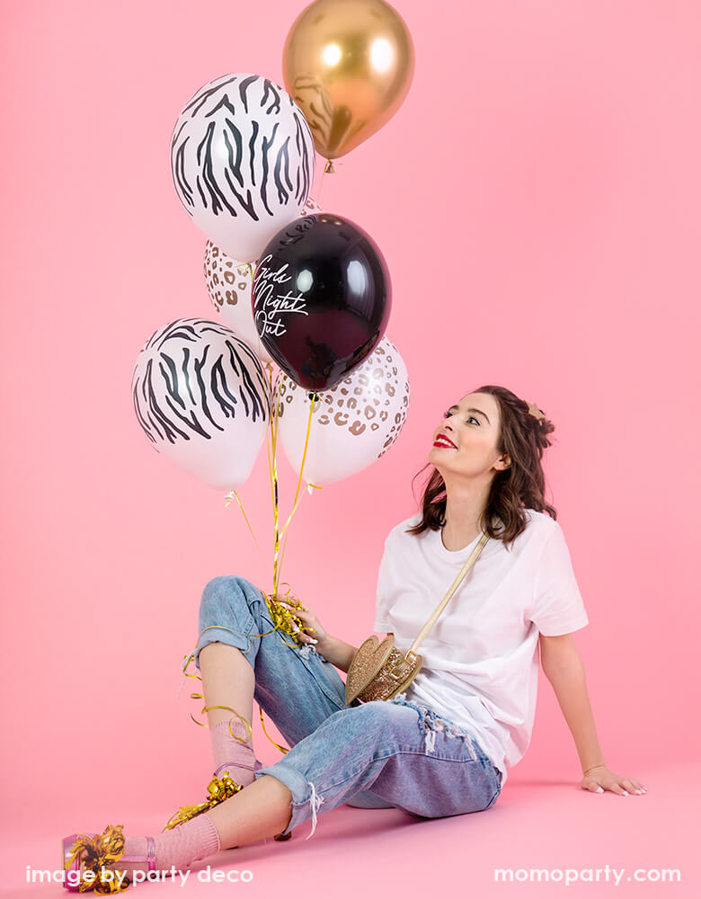 young lady sitting on a pink background, holding a balloon bouquet of Party Deco Leopard Print Latex Balloon, zebra print latex balloon, gold latex balloon, and black latex with "girls night out" text latex balloon