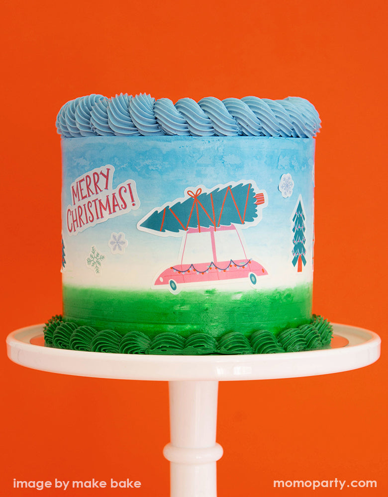 a blue white and green buttercream cake on a white cake stand, decorated with Make Bake shop Let's Get A Tree! Cakescape Edible Stickers with a truck carry christmas tree, snowflakes, "merry christmas" sign, many kinds of trees designs. These easy baking hacks cakescape edible stickers so easy to make decorating holiday cakes so much easier!