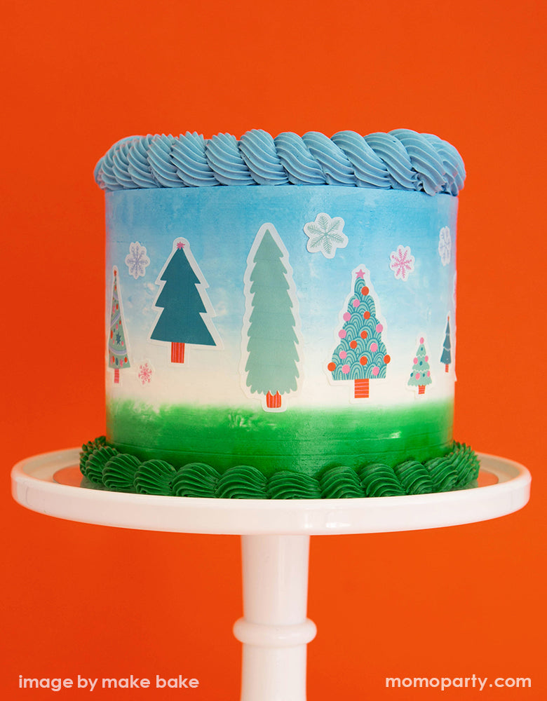 a blue white and green buttercream cake on a white cake stand, decorated with Make Bake shop Let's Get A Tree! Cakescape Edible Stickers with snowflakes and many kinds of trees designs. These easy baking hacks cakescape edible stickers so easy to make decorating holiday cakes so much easier!