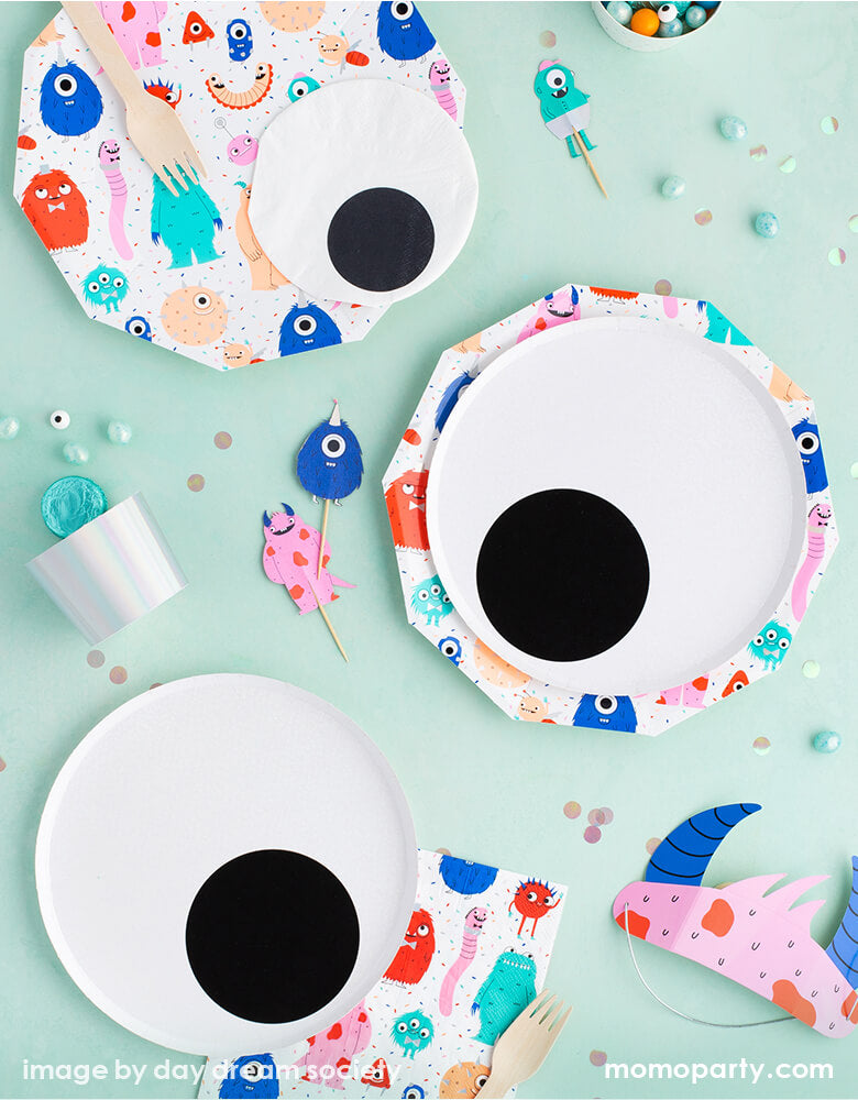 Little Monsters themed party table top view with Daydream Society Little Monsters Eyeball plates layered with Little Monsters Large Plates, with Little Monsters Large Napkins, Little Monsters Eyeball Small Napkins, Little Monsters Party Headband Set, silver baking cups with candies around, for a modern cute little monster themed birthday party, halloween party