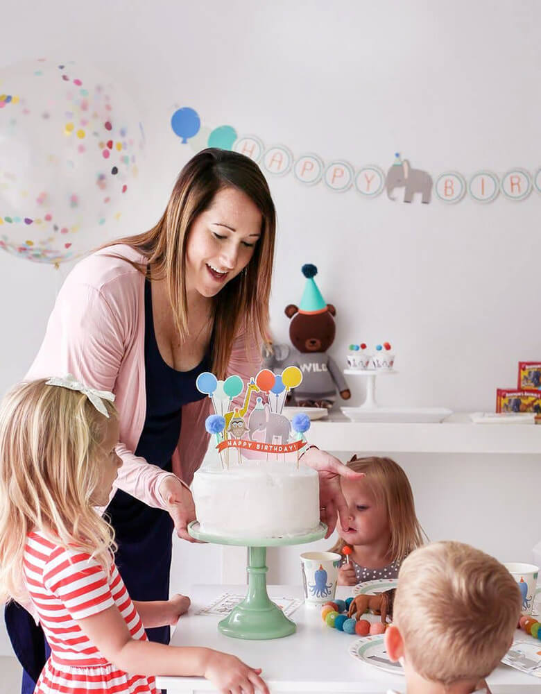 A mom holding a birthday cake for an animal themed birthday party for kids decorated with Lucy darling's party decorations including birthday banners on the wall, animal themed cake toppers and party tabletops