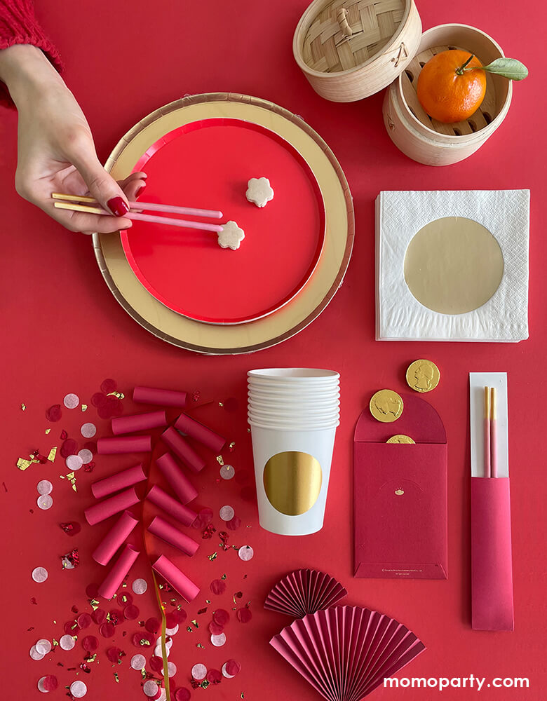 Momo Party's Modern Lunar-New-Year Party ideas with red small round plate overlap a GOLD LARGE PLATES, with a hand holding 2 strawberry flavor pocky sticks as chopstick, a gold dot cup, a red envelope with gold chocolate, gold dot napkin, a mandarin inside a mini Bamboo Steamer, paper rolls with confetti as firework. With all the gold and red that symbolizes happiness and prosperity over a red background, These modern and fun party supplies is new way to celebrate Lunar New Year!   