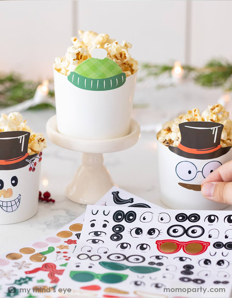 Holiday family baking activities with My mind's Eye - Make Your Own Snowman Food Cups. a kid holding a decor sticker putting on a white food cup with classic snowman black hat, there are a finished snowman food cup on the side, and a blank food cup with green sweater hat on a mini cake stand waiting to be decorated. Decorate them with your kids with the snowman stickers and have a fun-filled family time this holiday season. They're perfect for baking cupcakes right in the oven.