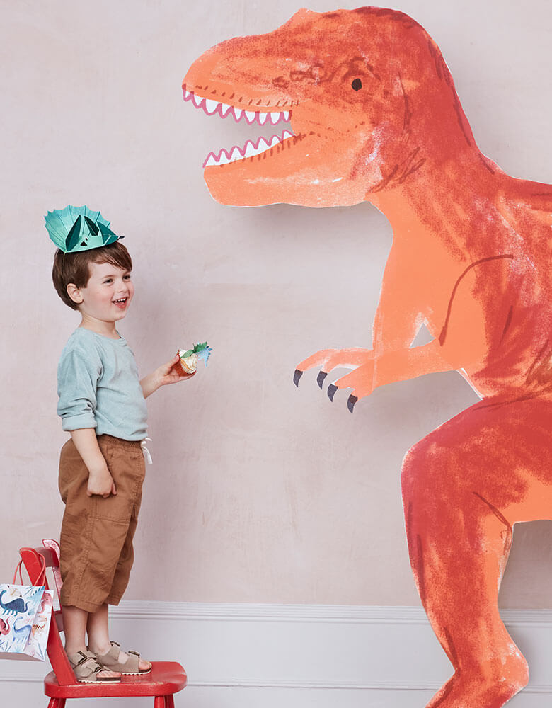 A boy in a dinosaur themed party wearing a dinosaur design hat and carries dinosaur party favor bag next to a giant t-rex cardboard standout