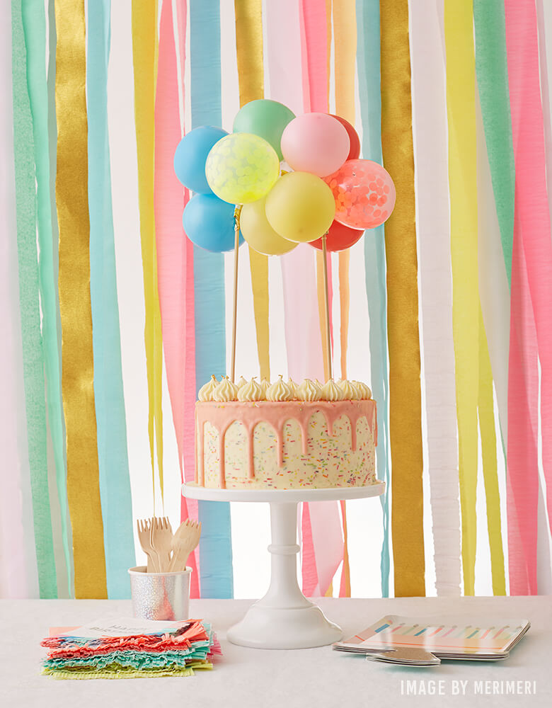 A birthday party dessert table with Meri Meri's happy birthday cake plates, napkin, sparkle birthday cake with rainbow colorful mini balloon garland as cake topper
