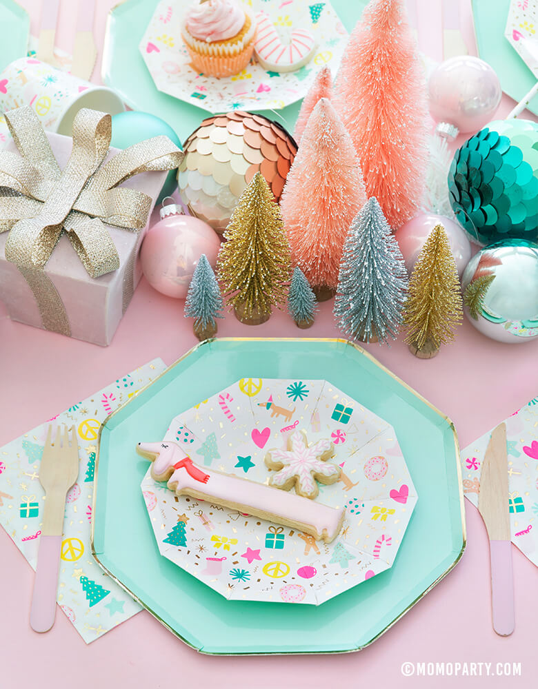 Merry and Bright Pastel Christmas table set up with cookies on top of Meri Meri Mint Large Dinner Plates and Daydream Society Merry and Bright Holiday Christmas Party Plates,Napkins and Cups, Pink wooden utensils, and gift box, Christmas Ornaments, mini pastel Sisal Trees as centerpiece for a modern pastel Christmas party celebration