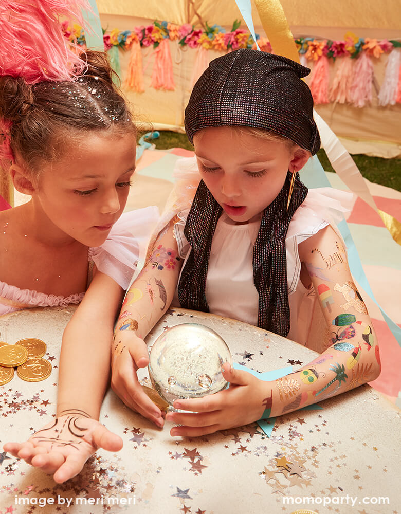 two girls dressed up like Gypsy Fortune Tellers, they watch a crystal ball  on a table, with full of cool bright colored and foil detailed temporary Tattoos on her arms