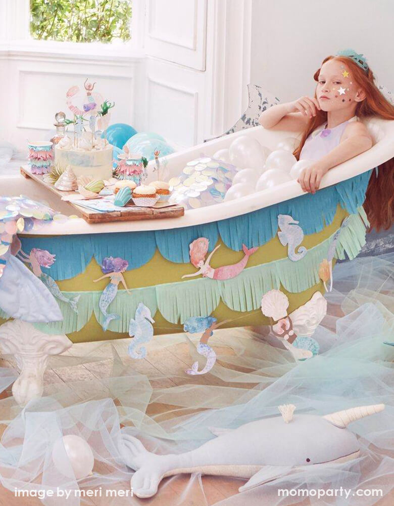 A Fun and gorgeous Mermaid Party at home. With a girl wearing a Mermaid Party Crown, shining stars sticker as face painting, she sitting inside bathtub like a mermaid, with lots of small latex balloons as bubbles. The bathtub decorated with Meri Meri Mermaid Fringe Garland, Light blue tulle fabric cover the ground as the ocean, with lots of desert and sweets on the bathtub caddy tray. What a creative fun dreamy mermaid party for a girls birthday, a mermaid lover, or a under-the-sea party