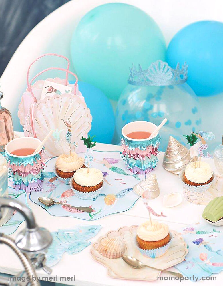 Detail look of a Modern Mermaid Party, There are Meri Meri Mermaid Swimming Plates, Watercolor Clam Shell Plates holding up cupcakes with Mermaid Cupcake topper, Seahorse Napkins, Mermaids Swimming Napkins, and Mermaid Scalloped Fringe Cups next to them, Mermaid seashell party bag with mermaid tattoo inside, sea shells as decorations. with Mixed blue,teal mint colored latex balloon behind it. What a fun dreamy mermaid party for eight kids birthday, or who love mermaid, or a under-the-sea party celebration