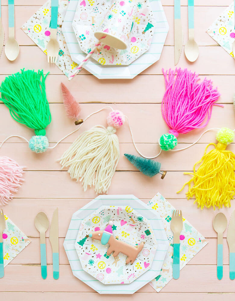 Daydream Society Merry and Bright Holiday Party Supplies on a pink table filled with festive colorful tassels and Christmas themed decorations 