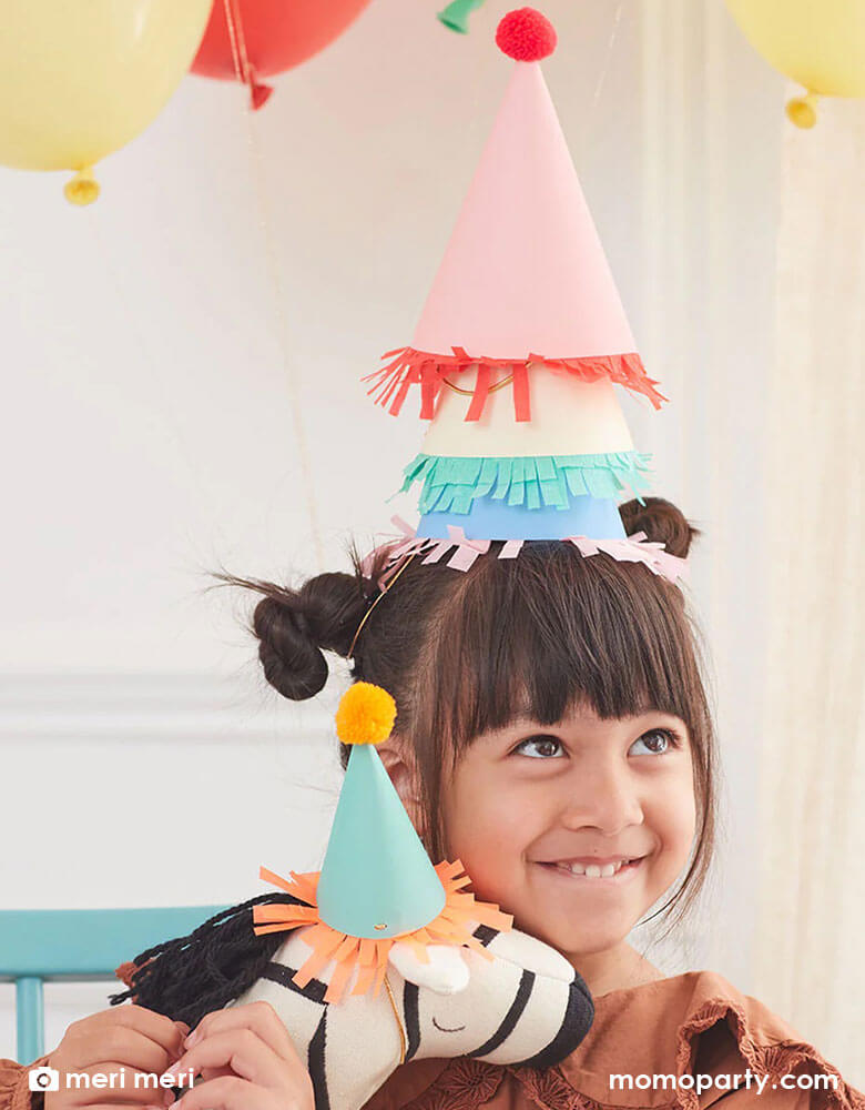 A girl wearing multiple party hats in bright colors holding a zebra stuffy what wears Momo Party's 2.25 x 4" mini party hat by Meri Meri.