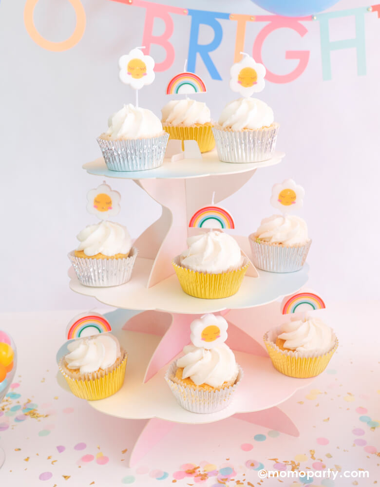 Cupcakes with Party Deco Rainbow and Daisy Birthday Candles as cupcakes toppers, on a Talking Tables Pastel rainbow We Heart Pastels Cake stand, with Meri Meri Multicolor Letter Garland Kit spells " To brighter Day" text behind it, and Cupcake Artisan Confettis on the table, for a Good vibes birthday party, a rainbow themed birthday, "Happy Day" themed celebration! 