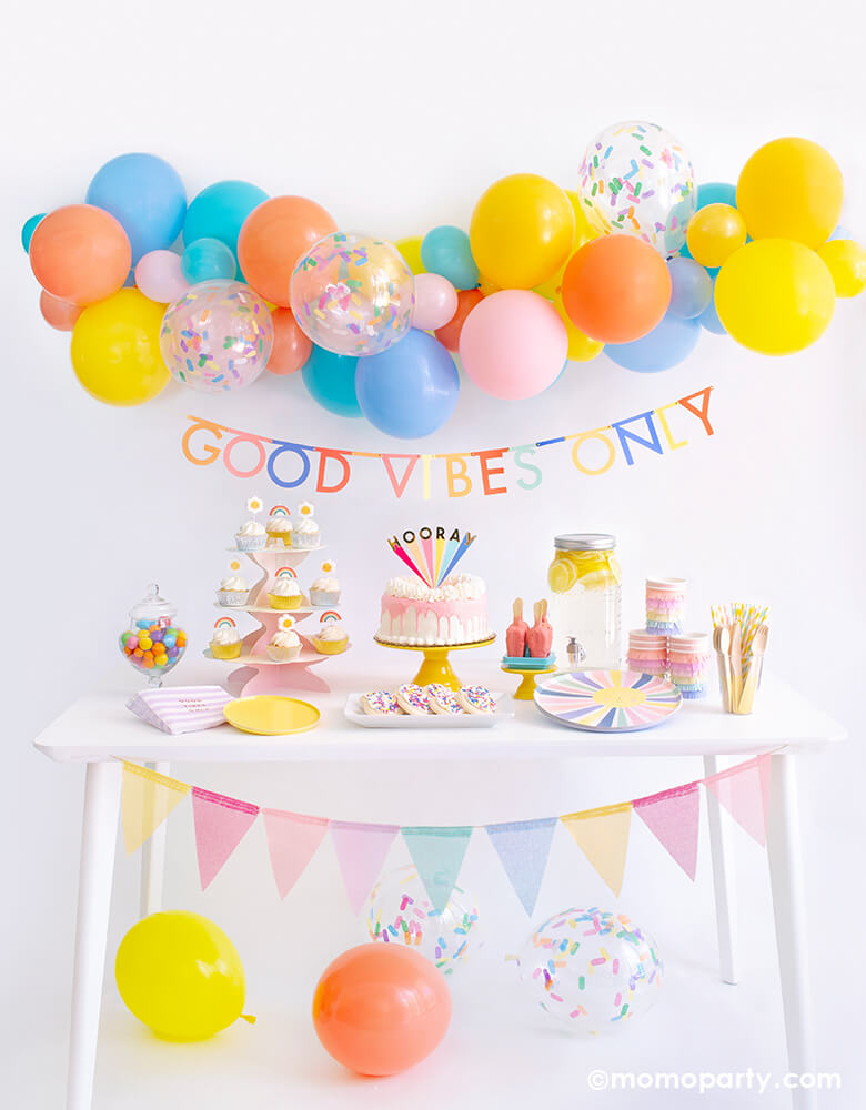 Momo party Good Vibes Only Party Box Kit. Included Meri Meri Rainbow Sun Dinner Plates, Oh Happy Day Yellow Side Plates, Meri Meri Rainbow Sun Fringe Cups, Party Deco Good Vibes Napkins, Yellow Wooden Cutlery, Mixed Pastel Striped Straws, cupcake with rainbow and daisy candles. Meri Meri Hooray cake topper on a dripping cake, colorful gumballs, Lemonade on the table. Meri Meri Multicolor Bunting Banner spelled as "good vibes only" text , Good Vibes 6-foot- Balloon Garland Cloud as decoration.