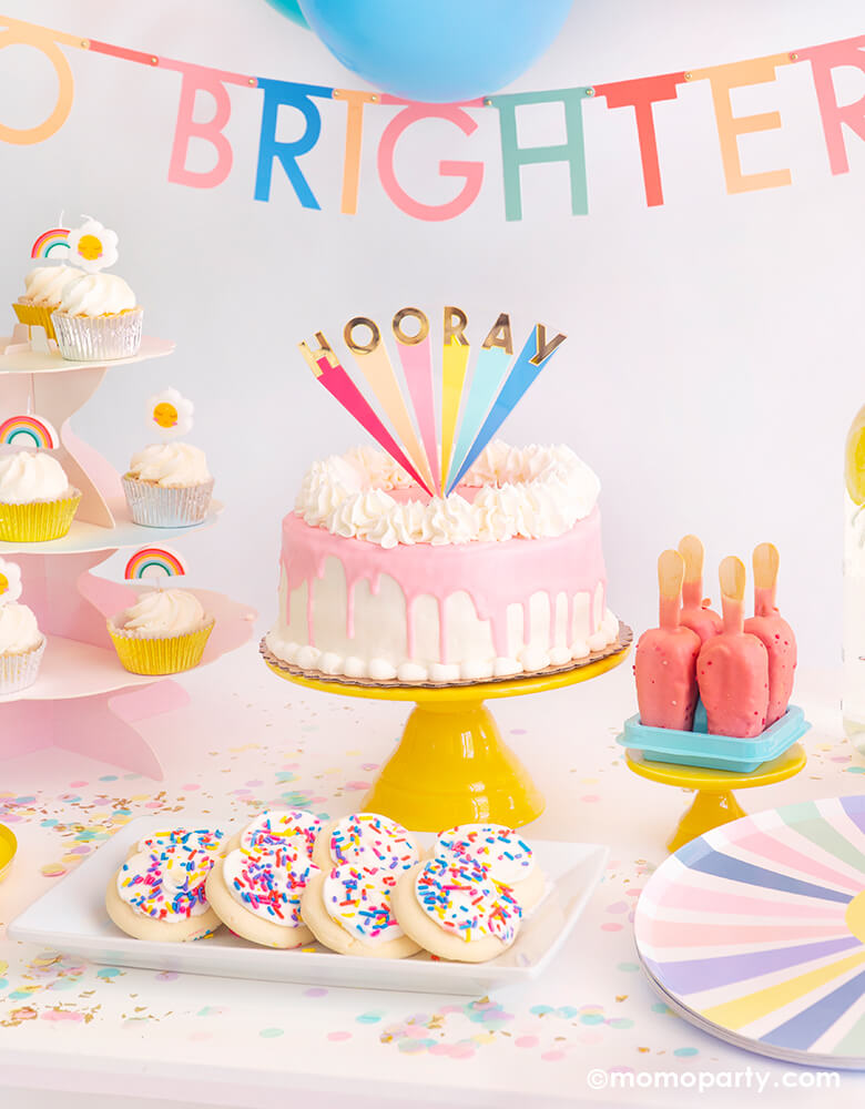 Party table close up look of modern dripping butter cream with Meri Meri Hooray Acrylic Cake Topper on a bright yellow cake stand, there cookies in the tray, cupcakes with daisy and rainbow mini candles on a pink cake stand. A neon colorful letter garland handing on top of th table
