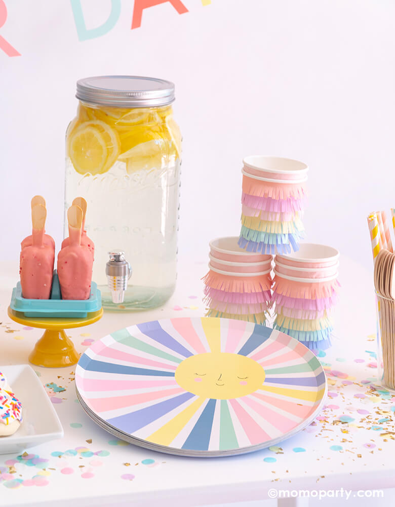 A Good Vibes themed party table, filled with Meri Meri Rainbow Sun Dinner Plates, Rainbow Sun Fringe Cups, lemonade in a Mason water dispenser, Popsicle sweets on a mini yellow cake stand,  Cupcake Artisan Confettis spread on the table. These Pastel rainbow themed modern partyware are perfect for a Good vibes birthday party, a rainbow themed birthday, "Happy Day" themed celebration!