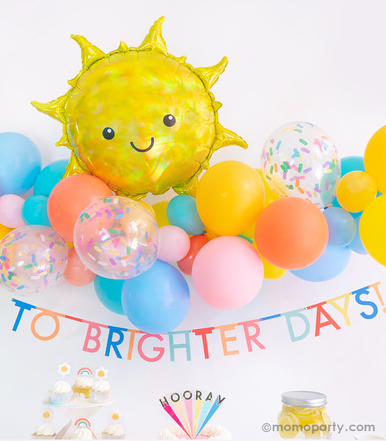 Momo party Good Vibes Only Party Box Look, Backdrop details of Anagram Iridescent Sun Foil Mylar Balloon over a colorful blue, yellow, coral, goldenode mixed latex balloon garland with confetti latex balloons, and Meri Meri Multicolor Bunting Banner spelled as "To Brighter Days!" text