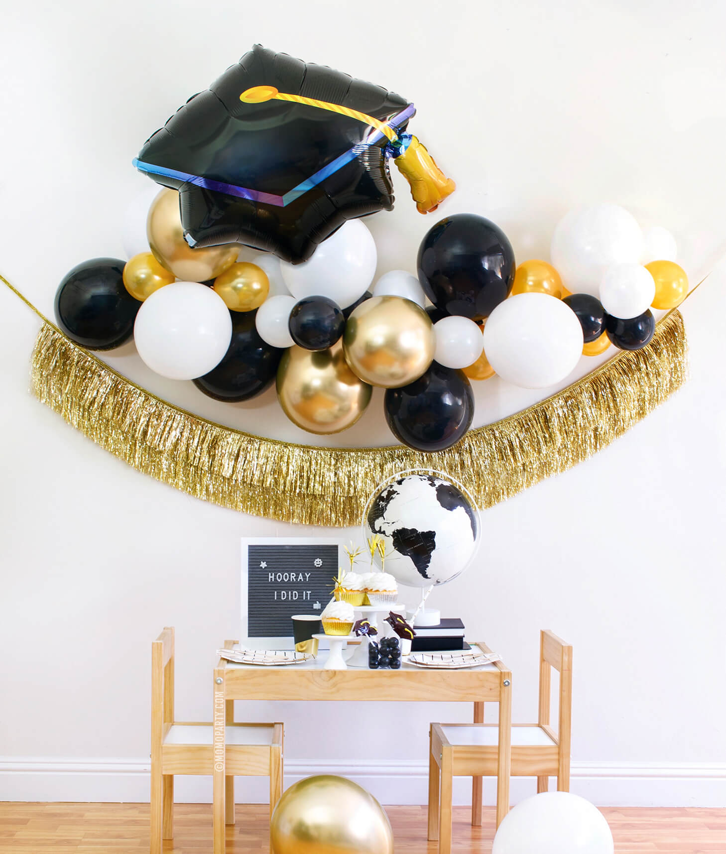 Momo party modern graduation party at home box, come with black, gold and white Latex balloon garland, Black Graduation Cap Foil Balloon, Gold Tinsel Fringe Garland as backdrop decoration. Black and Gold stripe paper plates, stripe napkins, Assorted Gold Dipped Cups, b&w globe, letter board with "hooray i did it" sign, cupcakes, black gum balls on a kid table for a graduation celebration