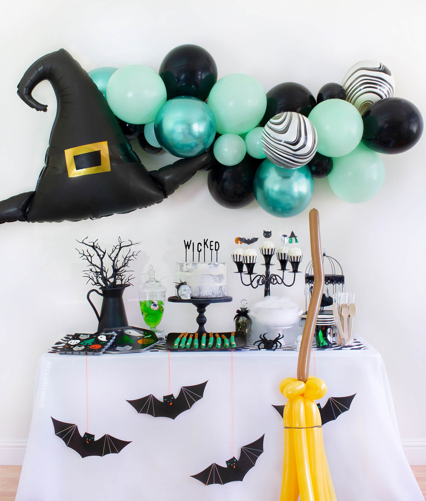 Momo Party 2020 Witch Please themed Halloween Party at home inspiration look with Anagram Witch Hat Satin Foil Balloon and pastel Green toned Latex Balloon Garland for wall decoration. Meri Meri Halloween Motif Dinner Plates, napkins and cups, cake with letter board cake topper spelled of "Wicked", witch cauldron, Apple jelly with eyeball candy in a jar, witch fingers pretzels on the dessert table, Meri Meri Hanging Bats and balloon animal witch broomstick in front of the table